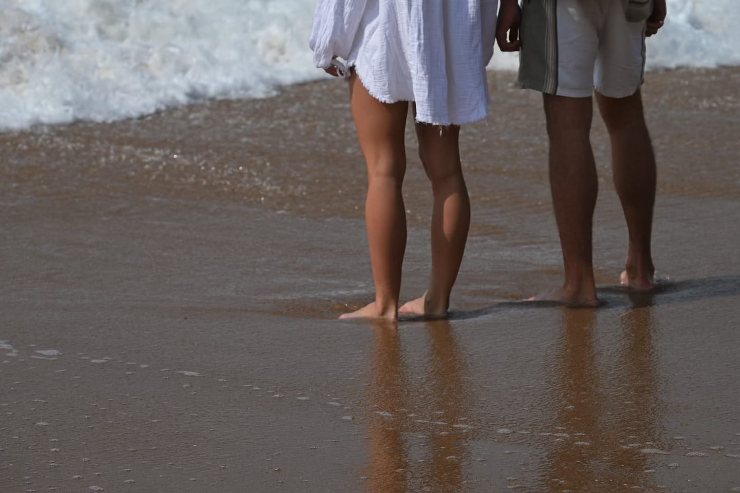 Two guests stand barefoot at the water’s edge as a wave washes around their feet