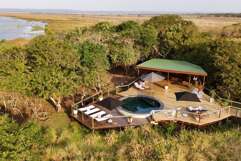 Aerial of the pool deck and green-roofed lounge among trees, the lake glinting beyond
