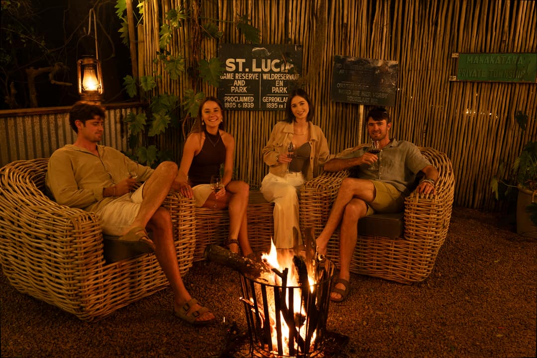 Two couples with glasses of wine sit in wicker armchairs around the fire at the boma.