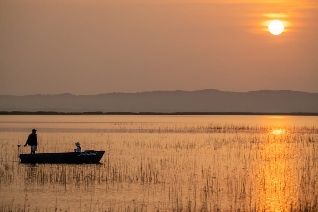 Two guests in the eco-boat at sunrise, one standing, the sun’s reflection streaking the water
