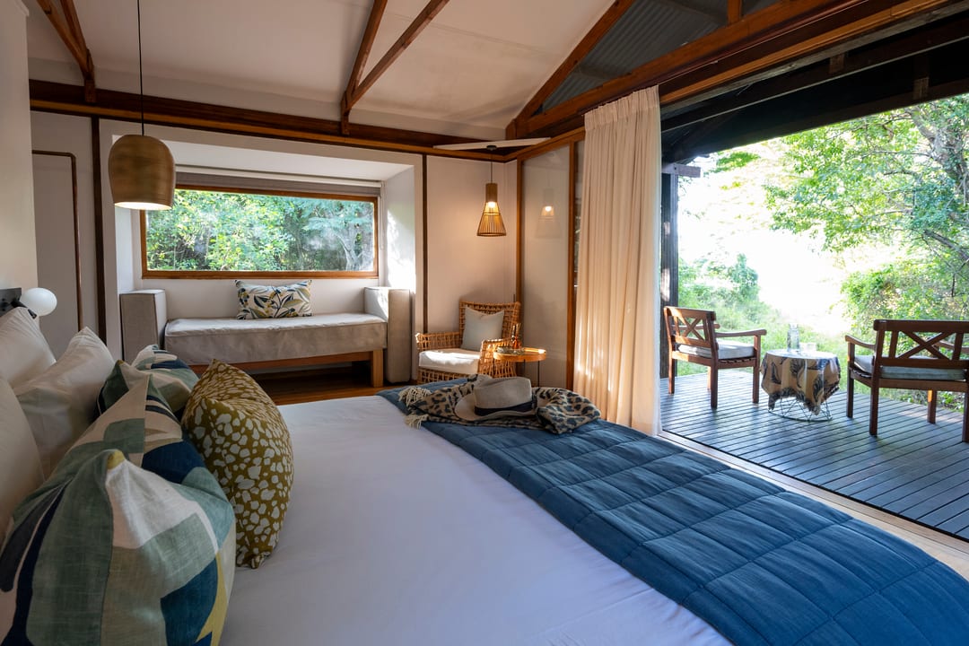 A bright suite in morning light, the bed facing a window seat and doors open to the deck.