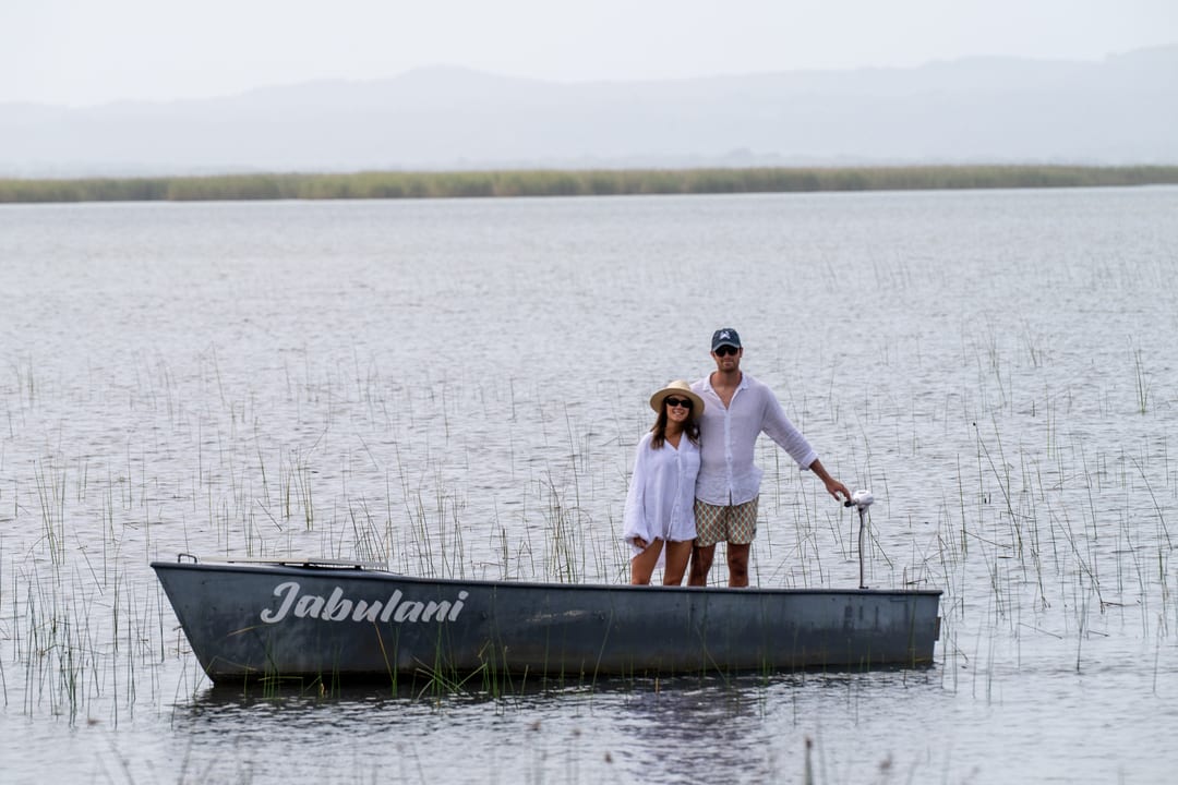A couple stand together in the electric skiff among reeds, hazy hills across the lake