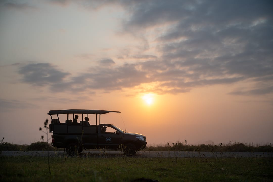 The game-drive vehicle in silhouette against a sunrise sky, two guests standing in the back
