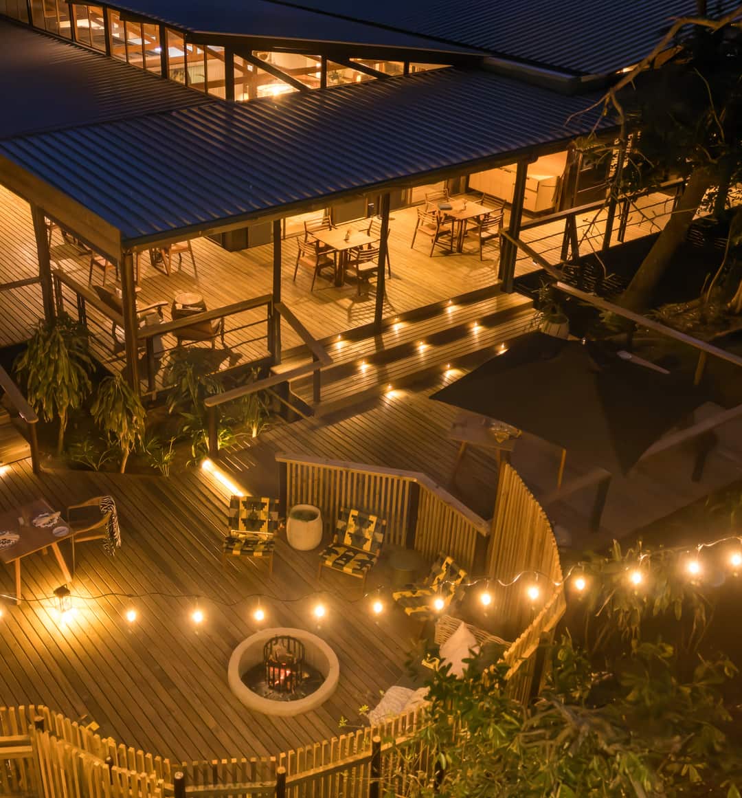 The main lodge from above at night, its dining tables lit beneath the roof and lit steps leading down to the deck boma, where the fire burns among string lights.