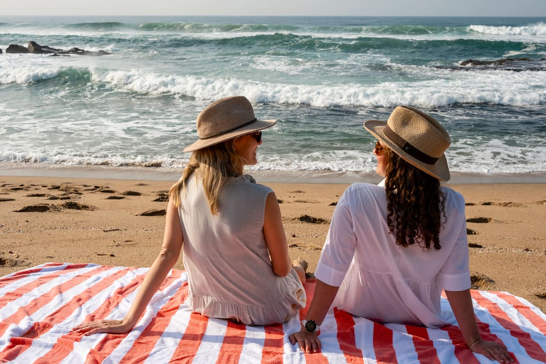 Two guests in straw hats sit on a striped towel and talk, surf and reef beyond