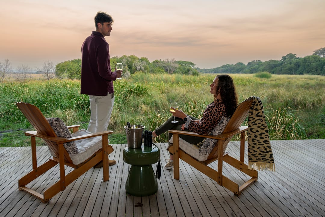 A couple with wine on the Honeymoon Suite deck at sunset, Adirondack chairs facing the wetland.