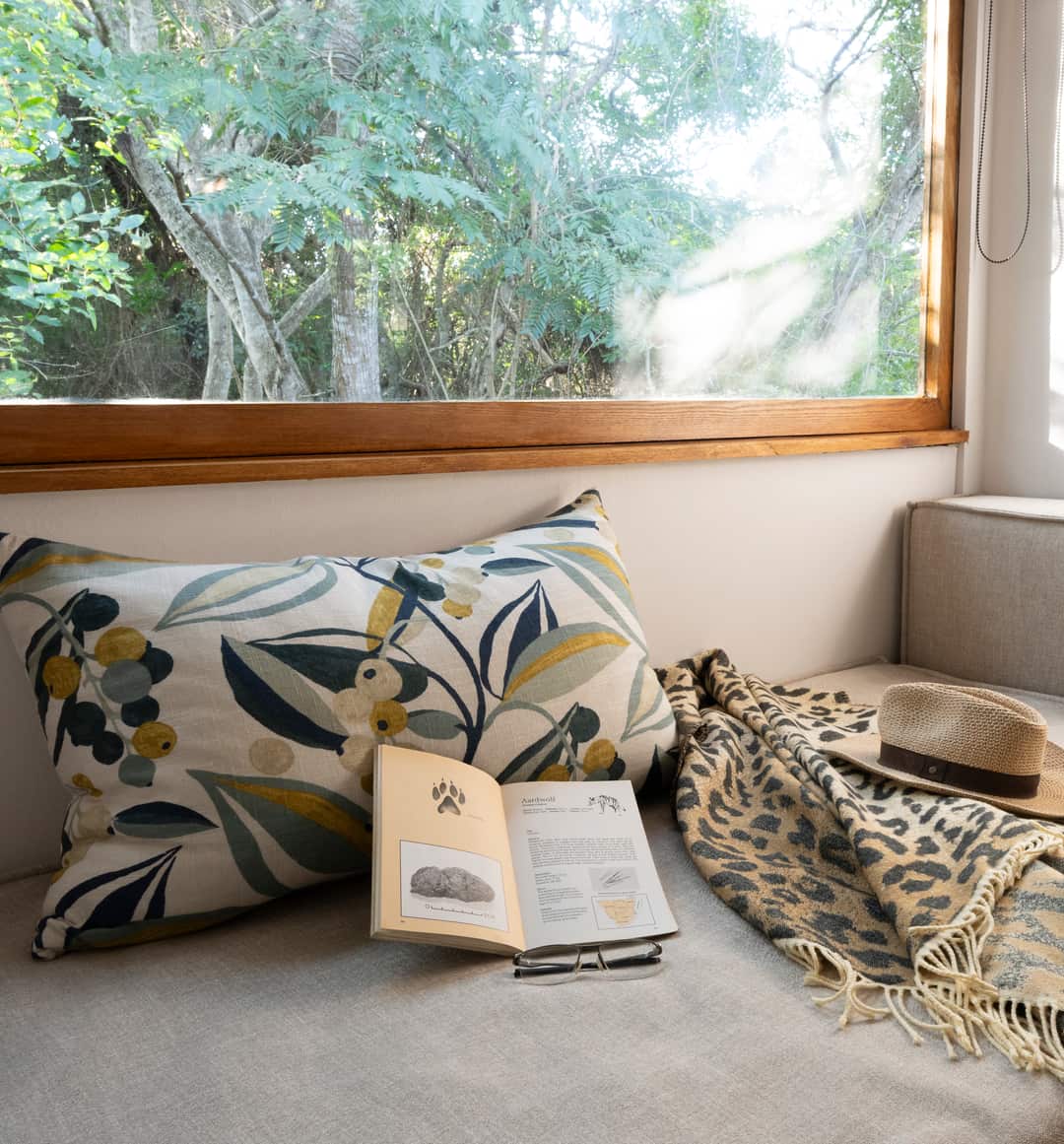 A window seat in a suite with a botanical cushion, an open wildlife guide and reading glasses, forest beyond.