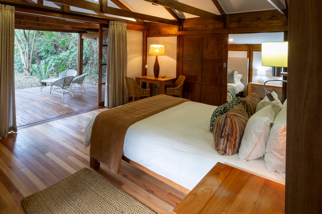 A suite bedroom in warm lamplight, doors open to a forest deck with chairs
