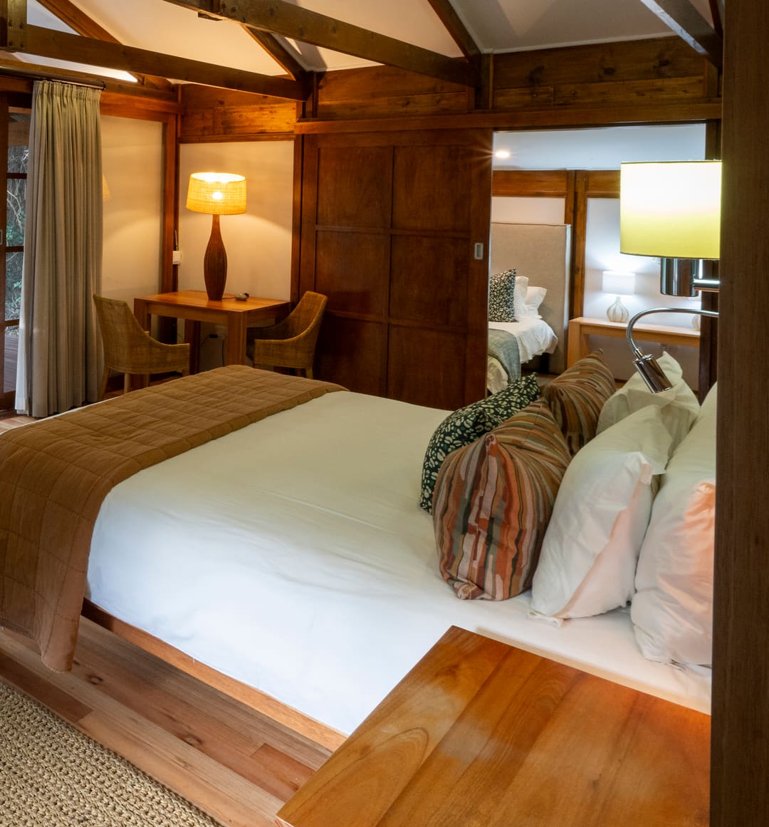 The Family Suite’s main bedroom with its lamps lit: the king bed beneath timber trusses, the second bedroom through the doorway and the doors open to the deck.