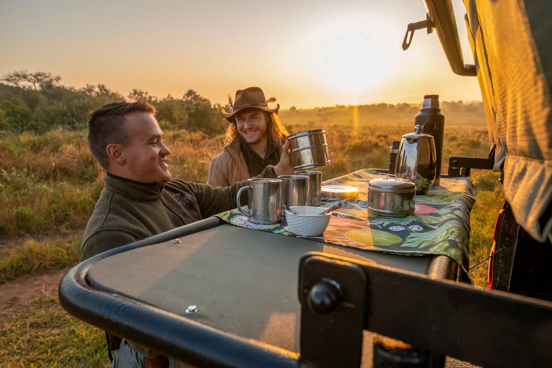 The guide reaches for a mug at a sunrise coffee stop by the tailgate, both laughing