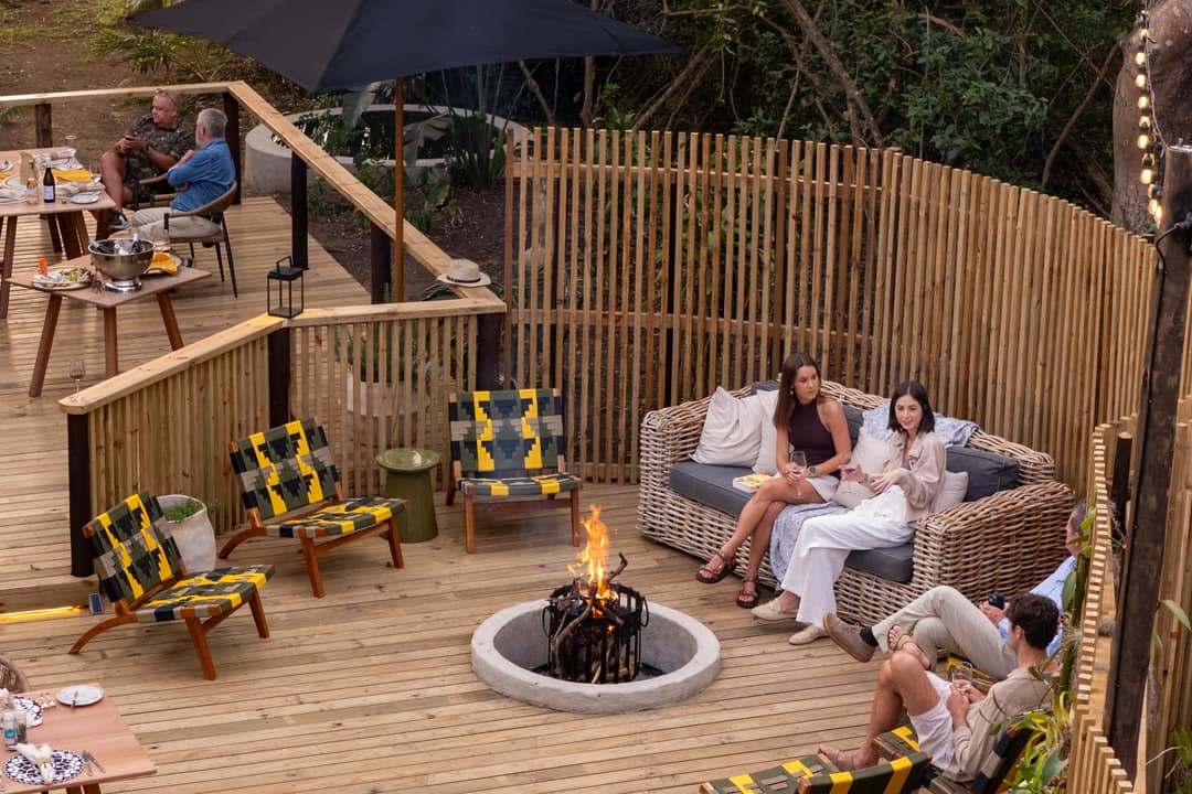 Guests relax on a rattan sofa beside the fire pit on the deck boma at dusk
