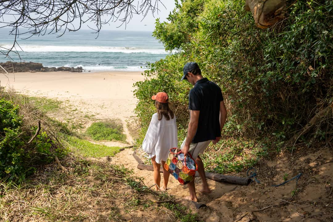 A couple follow a sandy path through coastal bush down to the beach, a basket in hand
