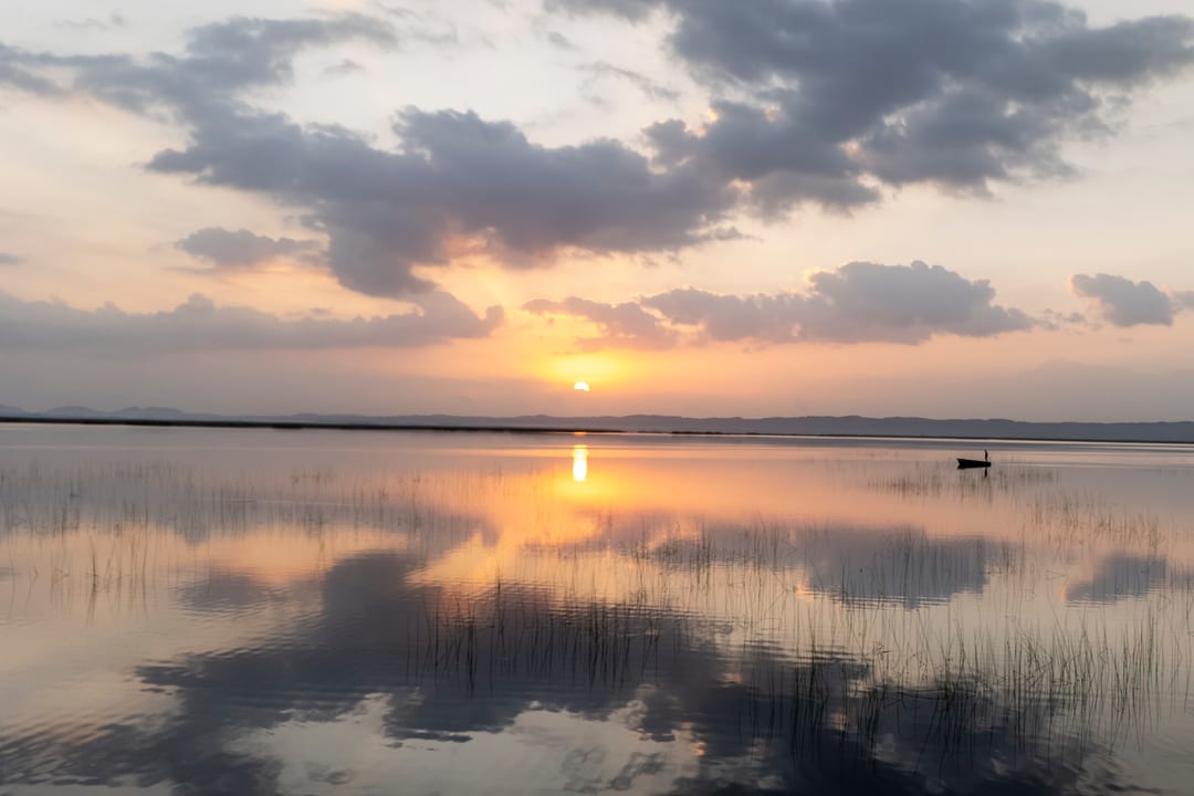 The sun sets through broken cloud over Lake St Lucia, clouds mirrored in the reedy shallows.