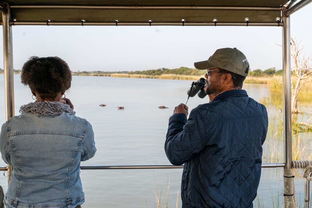 A couple at the boat rail, one with binoculars, watch three hippos surfacing on the lake