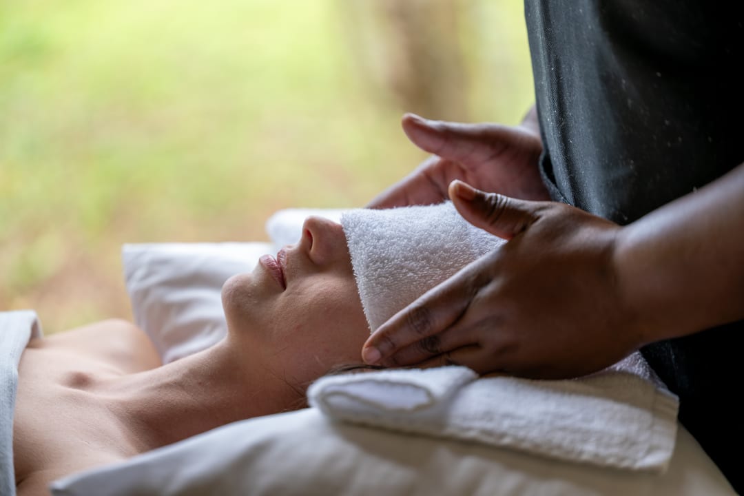 A therapist’s hands rest on a guest’s head during a treatment, a towel over her eyes