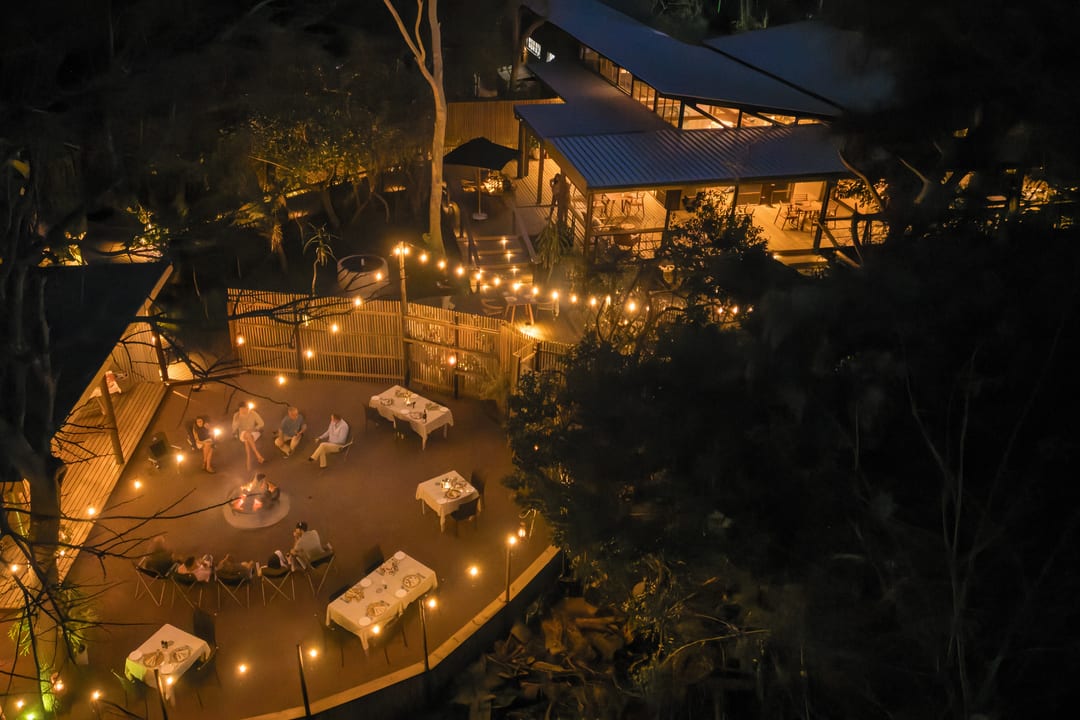The main boma glows with string lights and a fire at night, the lit lodge beyond.