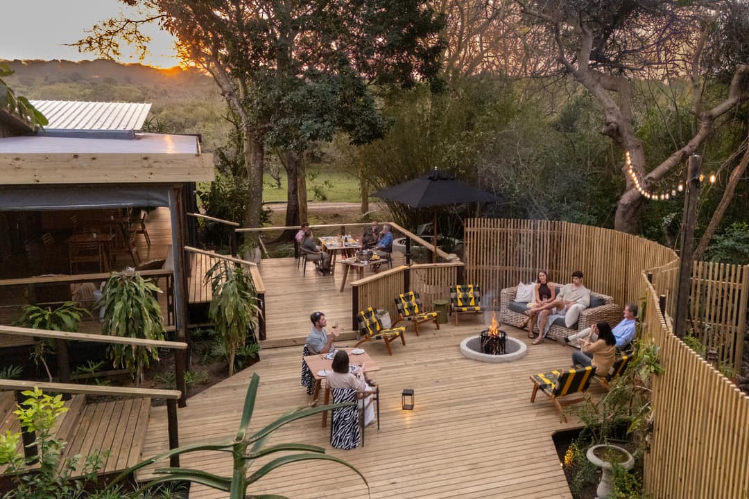 Guests gather around the fire on the deck boma at sunset, lodge roof and lake beyond