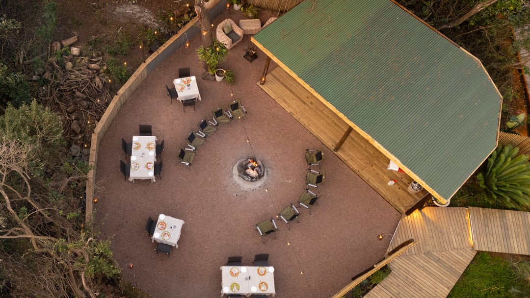 The main boma from directly above at dusk, its tables set around the fire pit beside the green-roofed pavilion and the lit boardwalk.