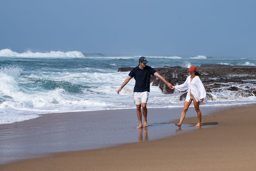 A couple hold hands at the water’s edge, spray rising off the rock shelves behind