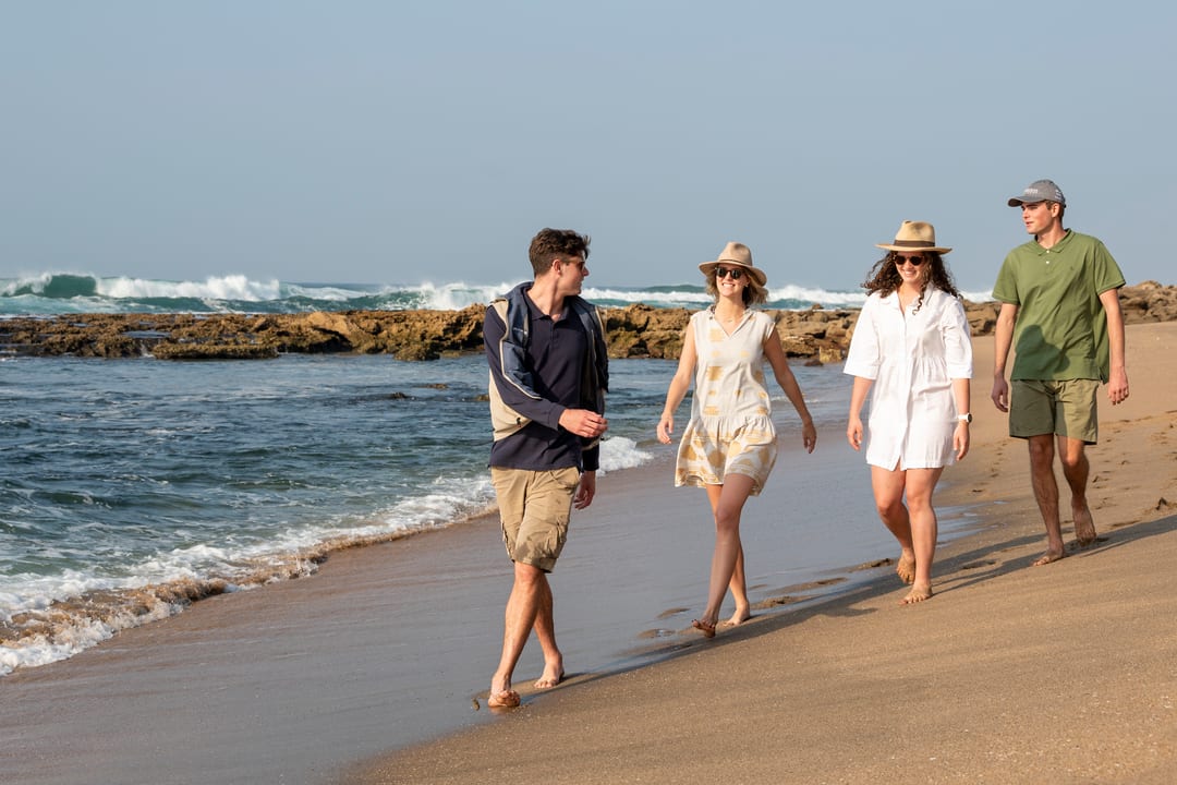 Four guests walk the tideline in bright morning light, reef and surf behind