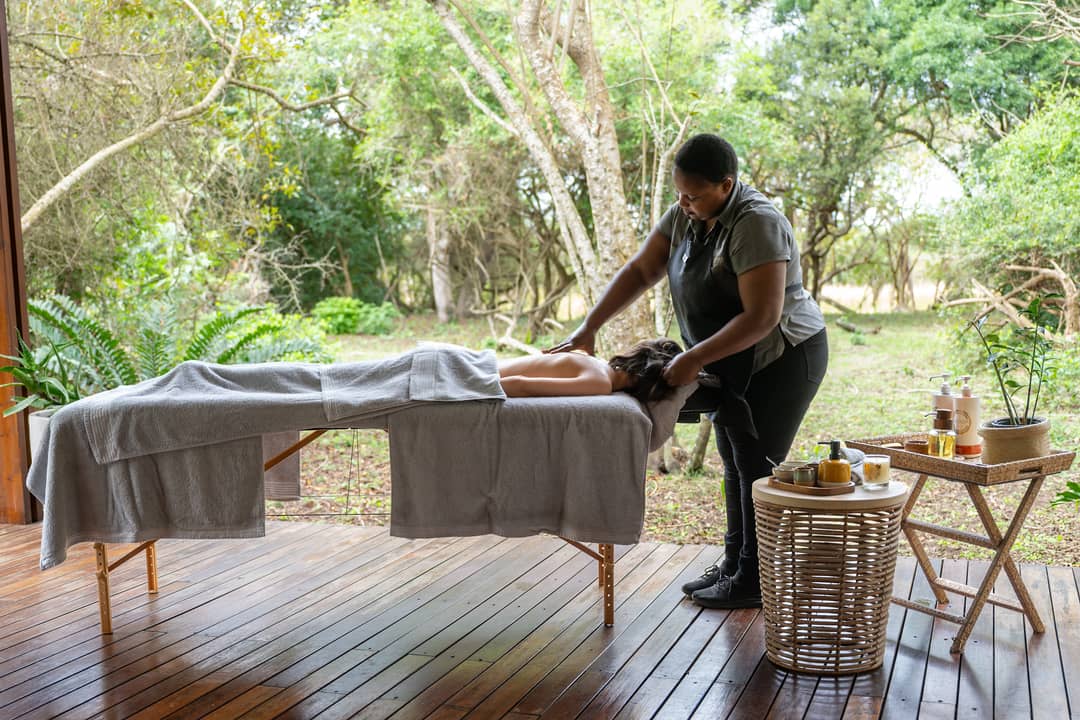 A therapist gives a back massage on an open timber deck among trees and ferns