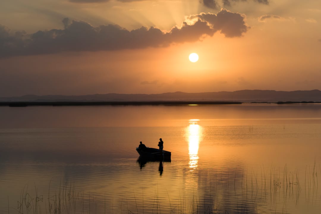 Two guests stand in an eco-boat beneath a low sun, a molten path of light on the water