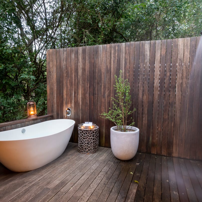 An outdoor bath and rain shower on a suite’s private deck at dusk, a lantern lit, forest behind