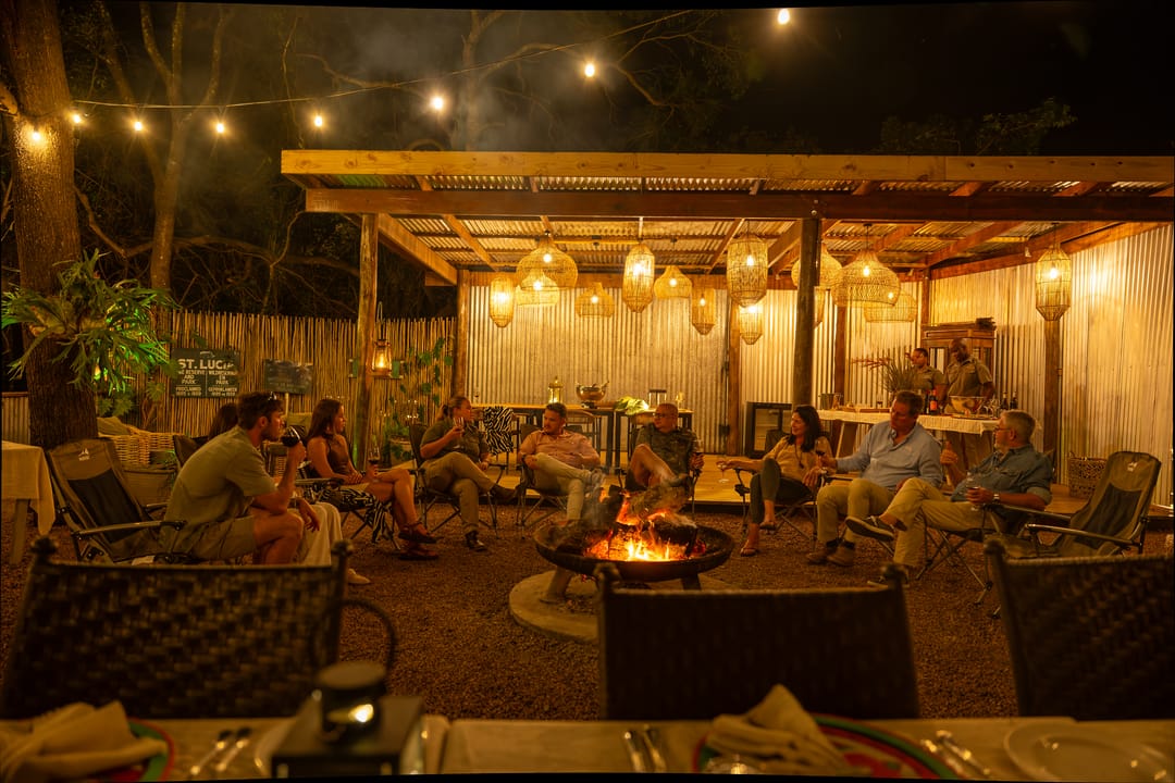 Guests in canvas chairs around the fire bowl in the main boma, lanterns lit in the pavilion