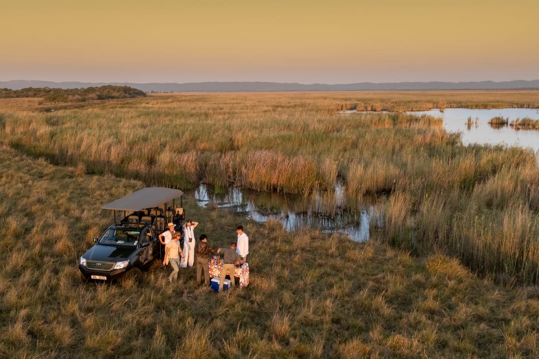 Seen from above at dusk, guests gather beside the vehicle for sundowners at the edge of the wetland.