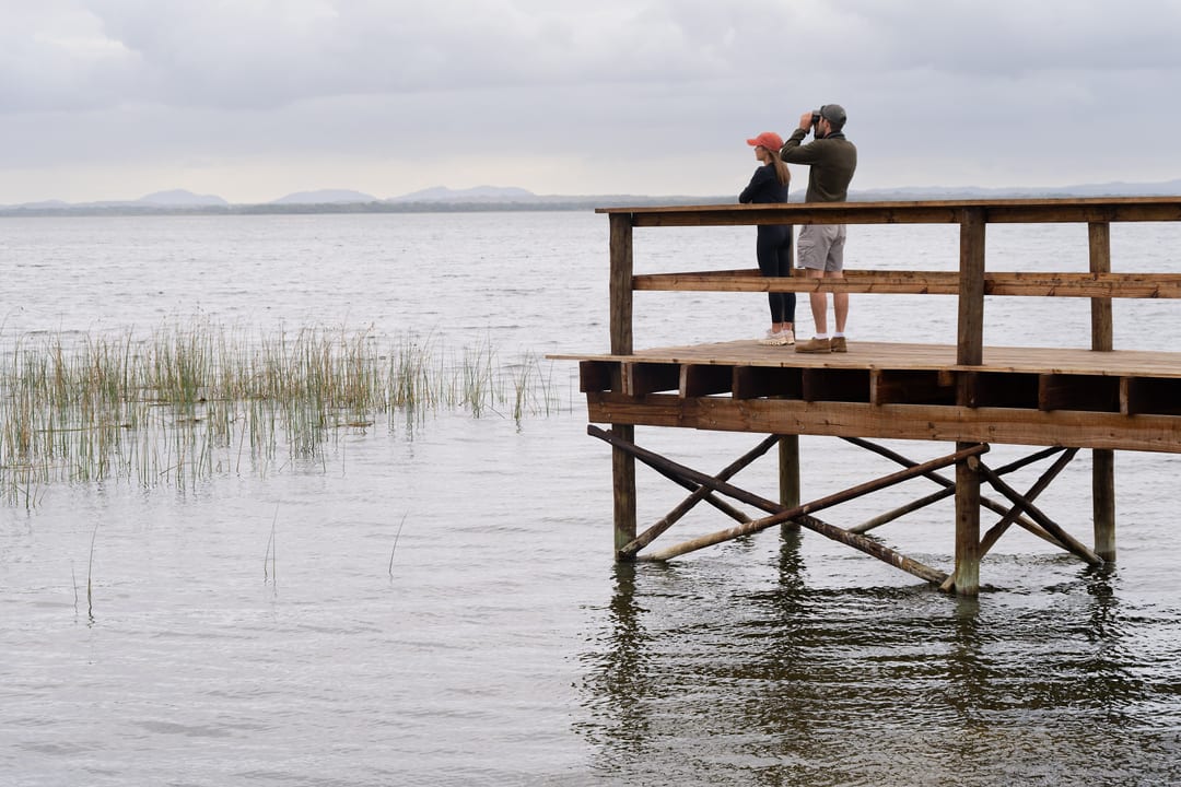 Two guests with binoculars on a raised timber deck over the lake, hills across the water