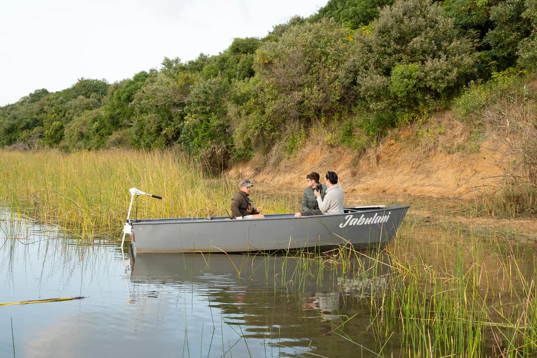 A guide and two guests sit in a skiff among reeds below a forested bank