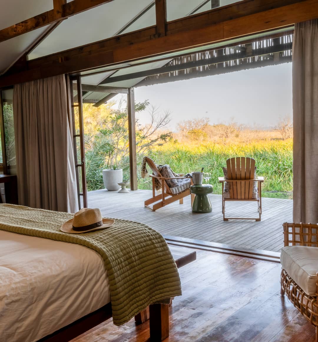 A suite bedroom in golden light, its walls opened onto a deck and sunlit wetland grasses.