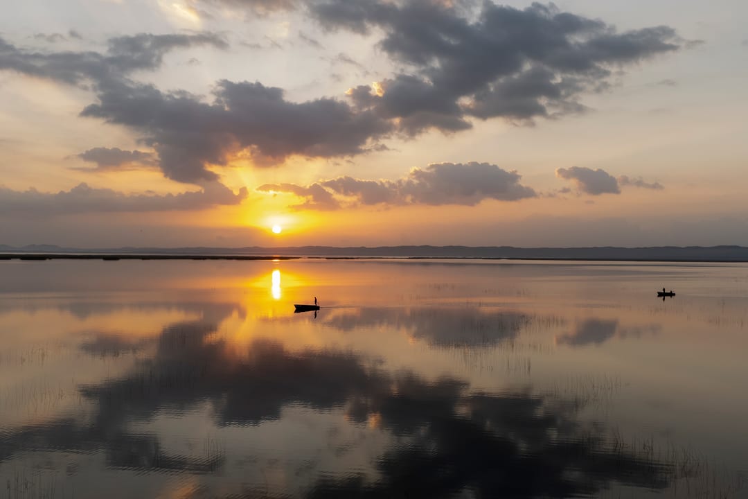 Sunrise breaks through dark cloud over a mirror-flat bay with two small boats