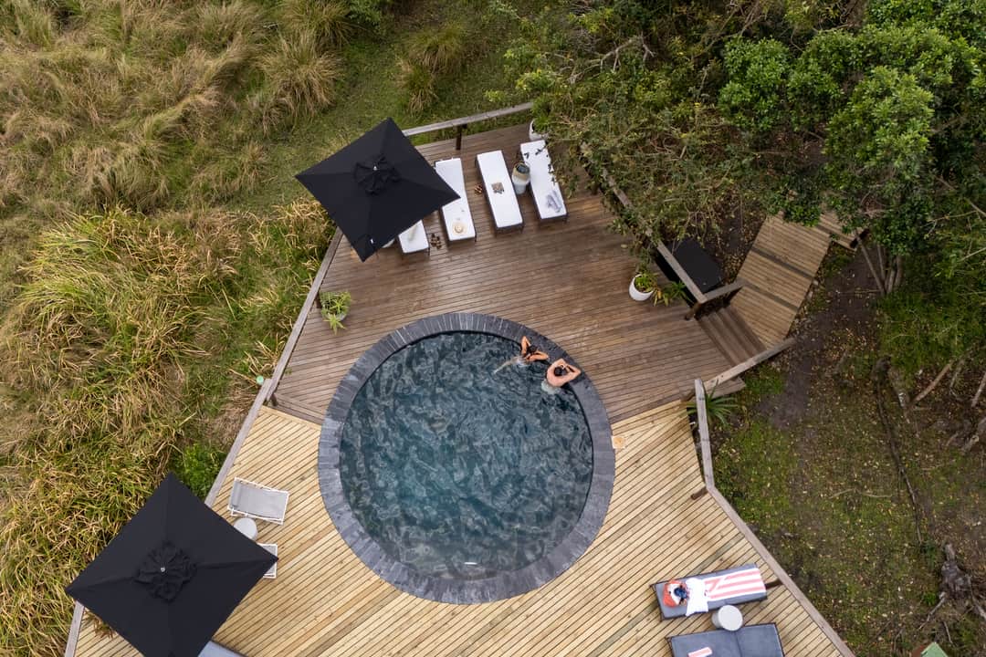 The round pool and its deck from directly above, two swimmers, black umbrellas and loungers.