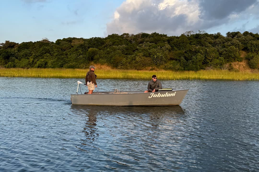 A guide steers a skiff with a seated guest across blue water below a forested shore