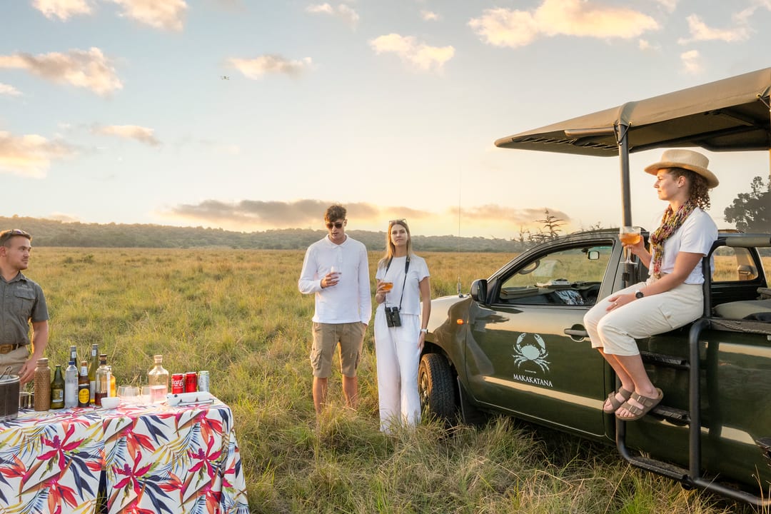 A guide pours drinks at a sundowner table while guests relax beside the vehicle