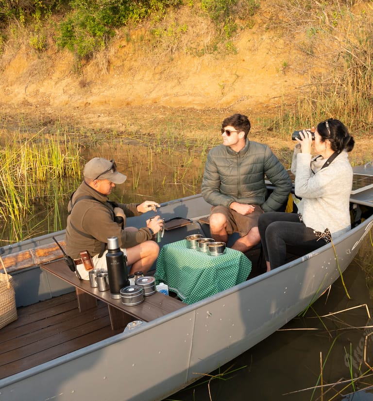 A guide makes coffee at the gingham-covered table on board an eco-boat while one guest looks out over the reeds and another scans the bank with binoculars.