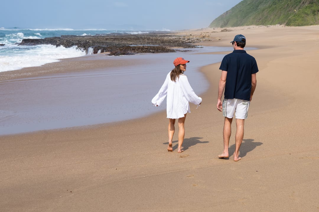 A couple walk barefoot on wet sand towards the rock shelves and a forested headland