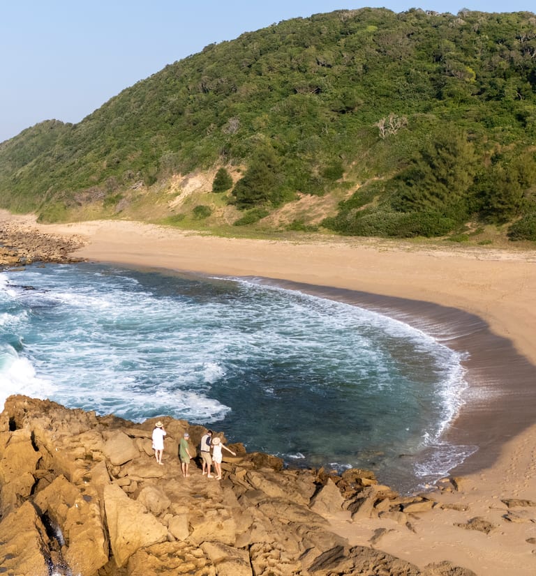 A curved, empty bay below forested dunes, with four guests small on the rocks at the water’s edge.
