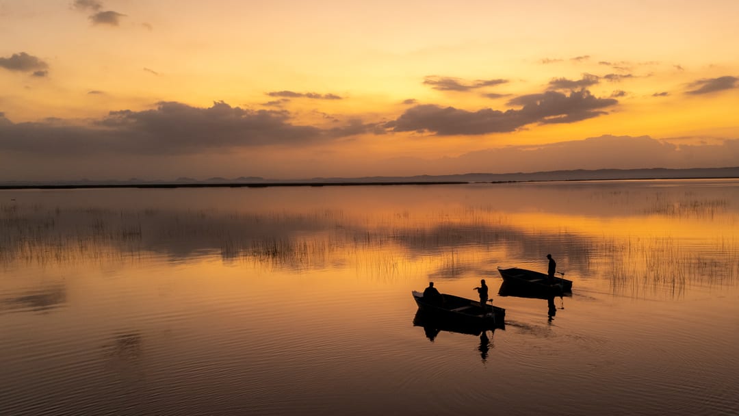 Aerial view of two eco-boats in silhouette on a mirror-calm lake at sunset, cloud reflected around them.