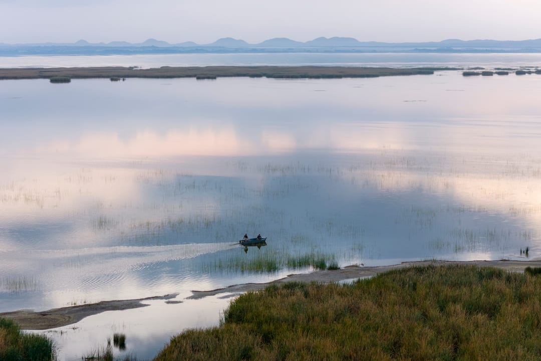A small boat crosses a vast pastel-blue lake at dawn, hills on the horizon