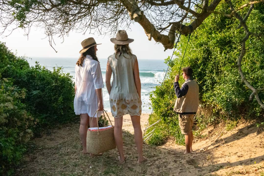 Guests with a picnic basket pause on the milkwood-framed dune path above the surf.