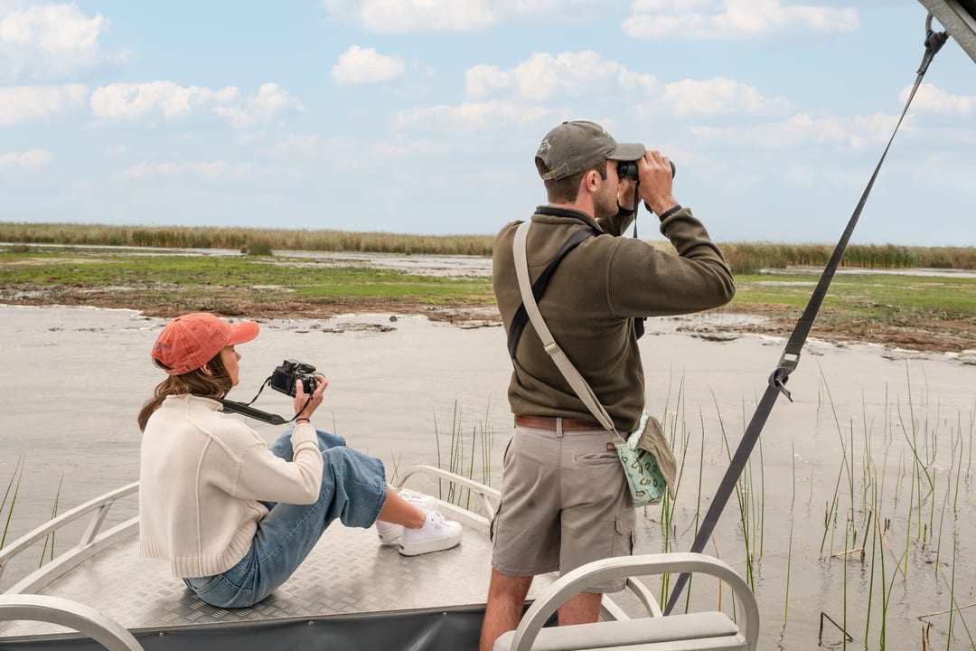 Two guests on the open bow, one with a camera and one with binoculars, scanning the reeds