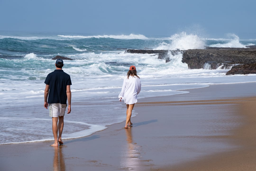 A couple walk barefoot towards the surf as a wave breaks over the rock shelf.