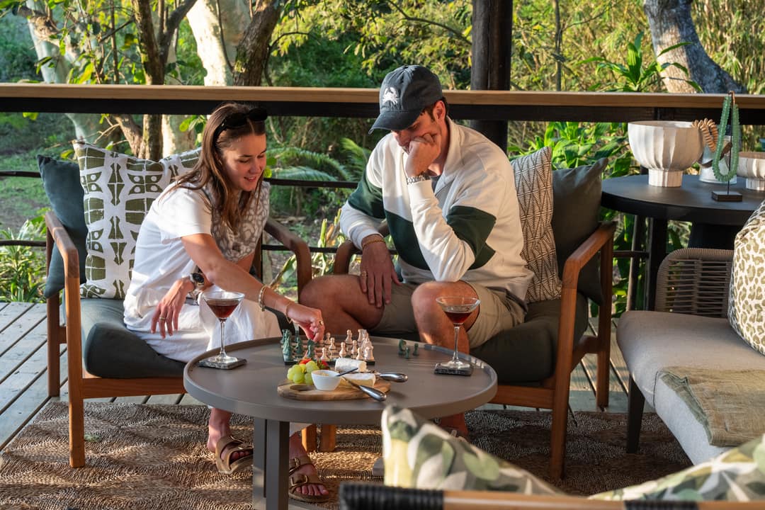 A couple laugh over a game of chess with drinks on a shaded veranda.