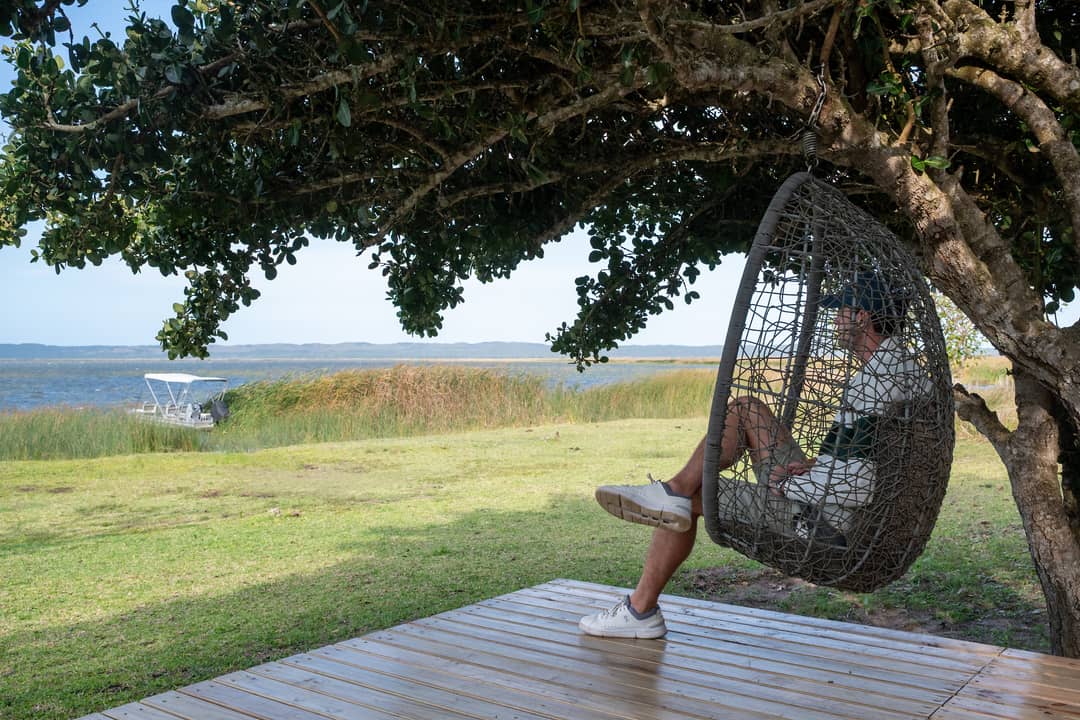 A guest relaxes in a hanging egg chair under a big tree, looking out to the lake