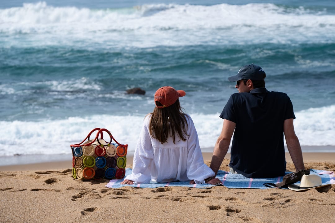 A couple sit on a beach blanket beside a woven basket, watching the surf roll in.