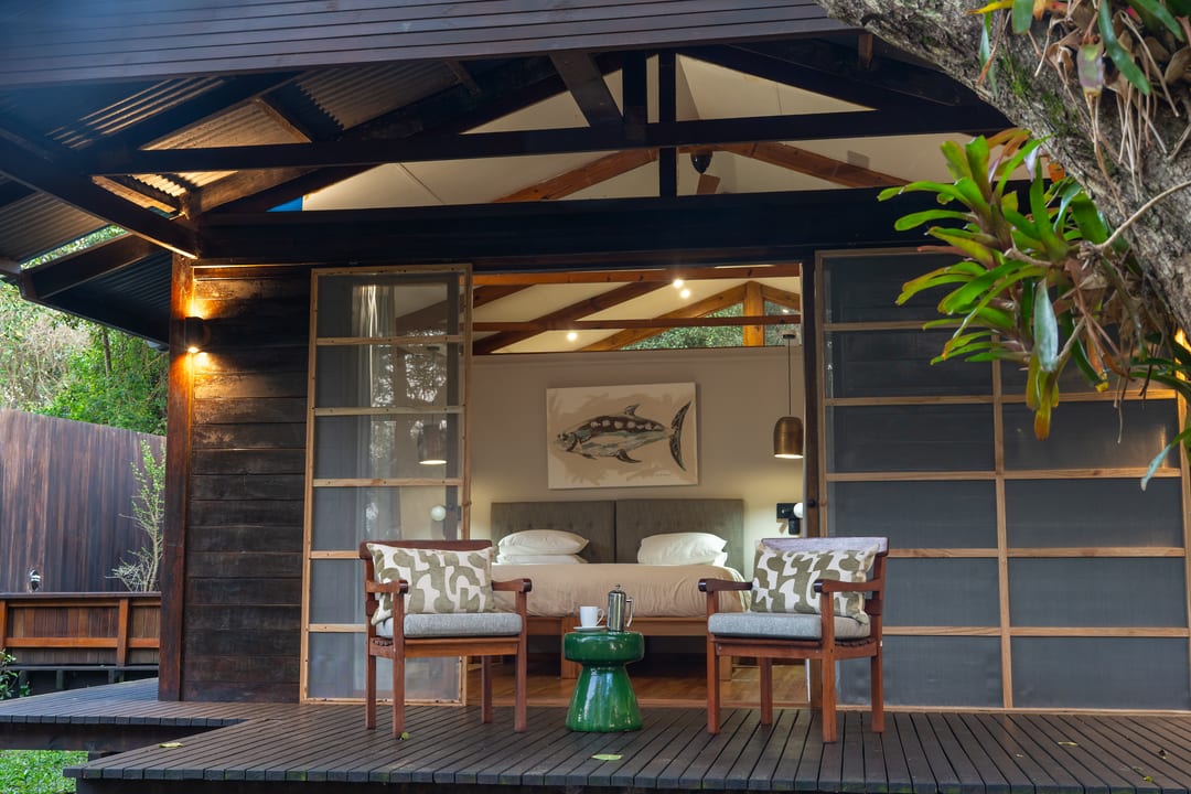 A dark timber suite at dusk, sliding doors open to a lit bedroom and two deck chairs