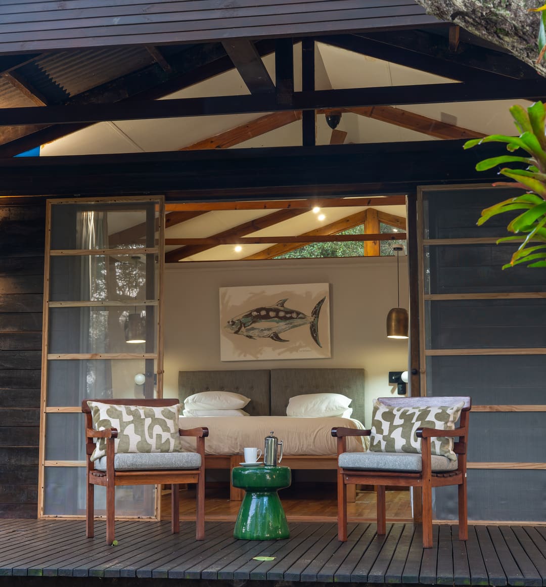 A suite at dusk, its sliding doors open from the lit bedroom onto a private deck with two armchairs.