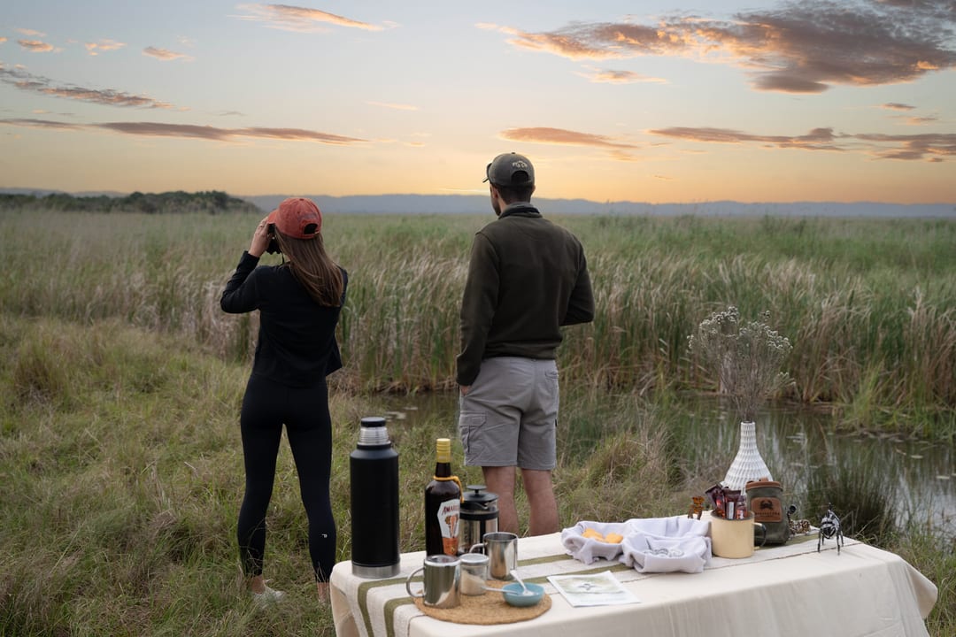 Two guests watch the low sun over the lake by a coffee table at the reed edge